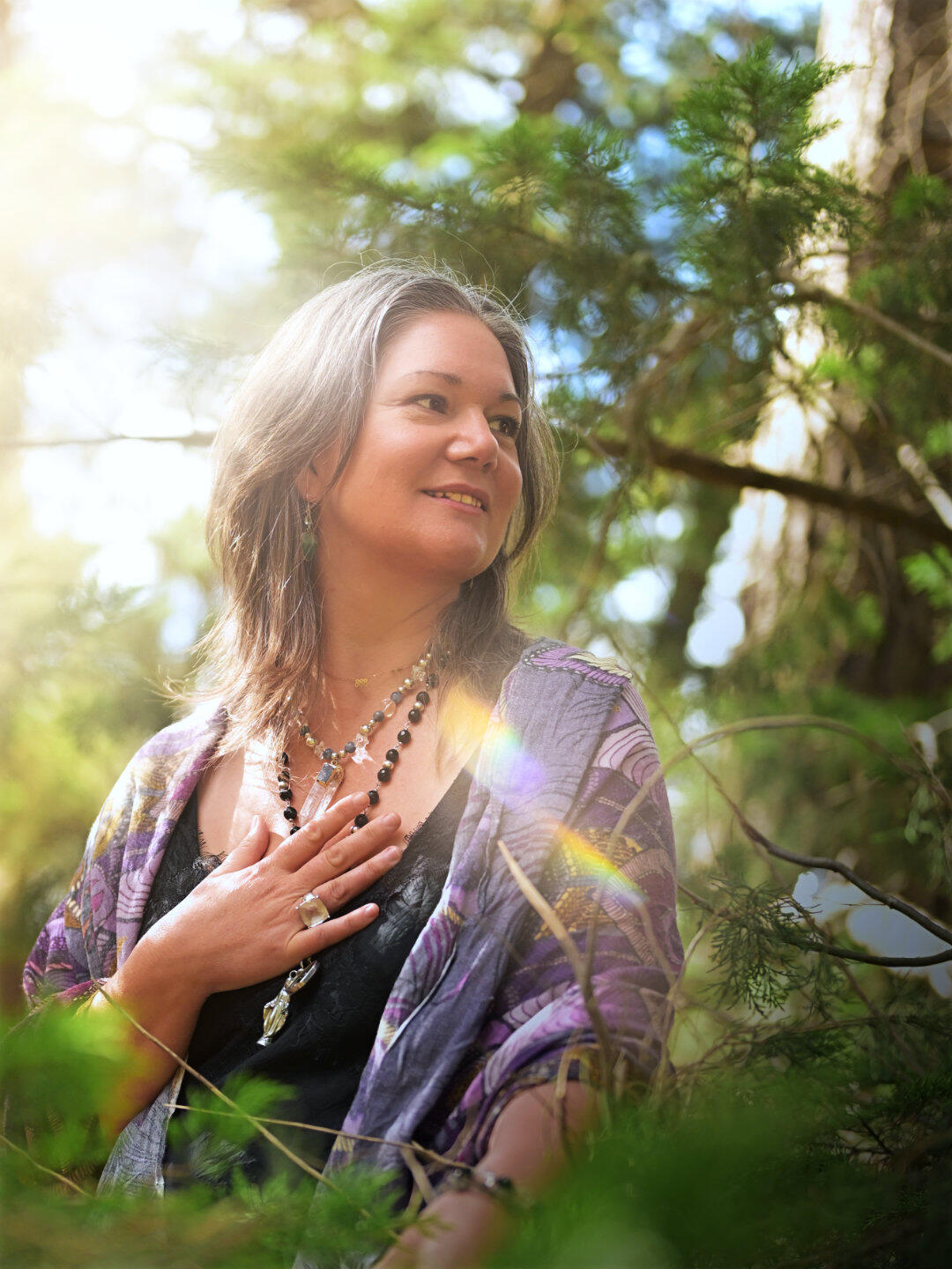 A woman in the forest in her priestess energy with hand to chest enjoying the glow of the afternoon sun streaming through the tree canopy.
